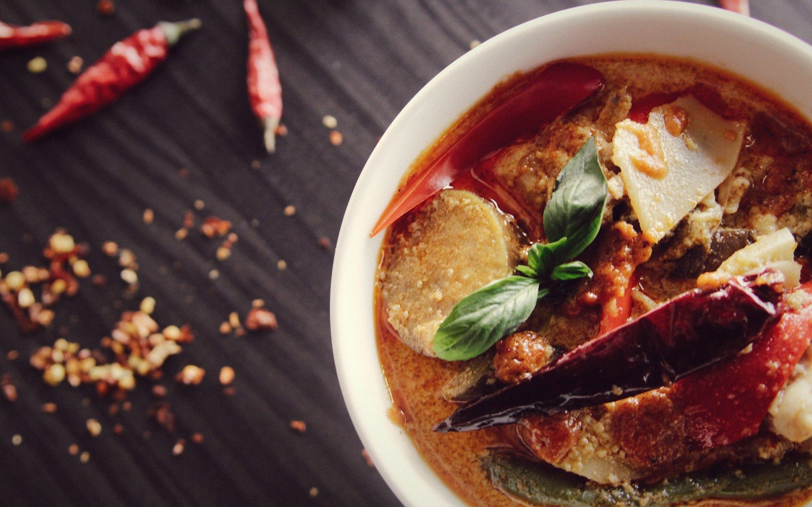 Bowl of red curry with dried chilies, basil leaves, and sliced vegetables on dark wood