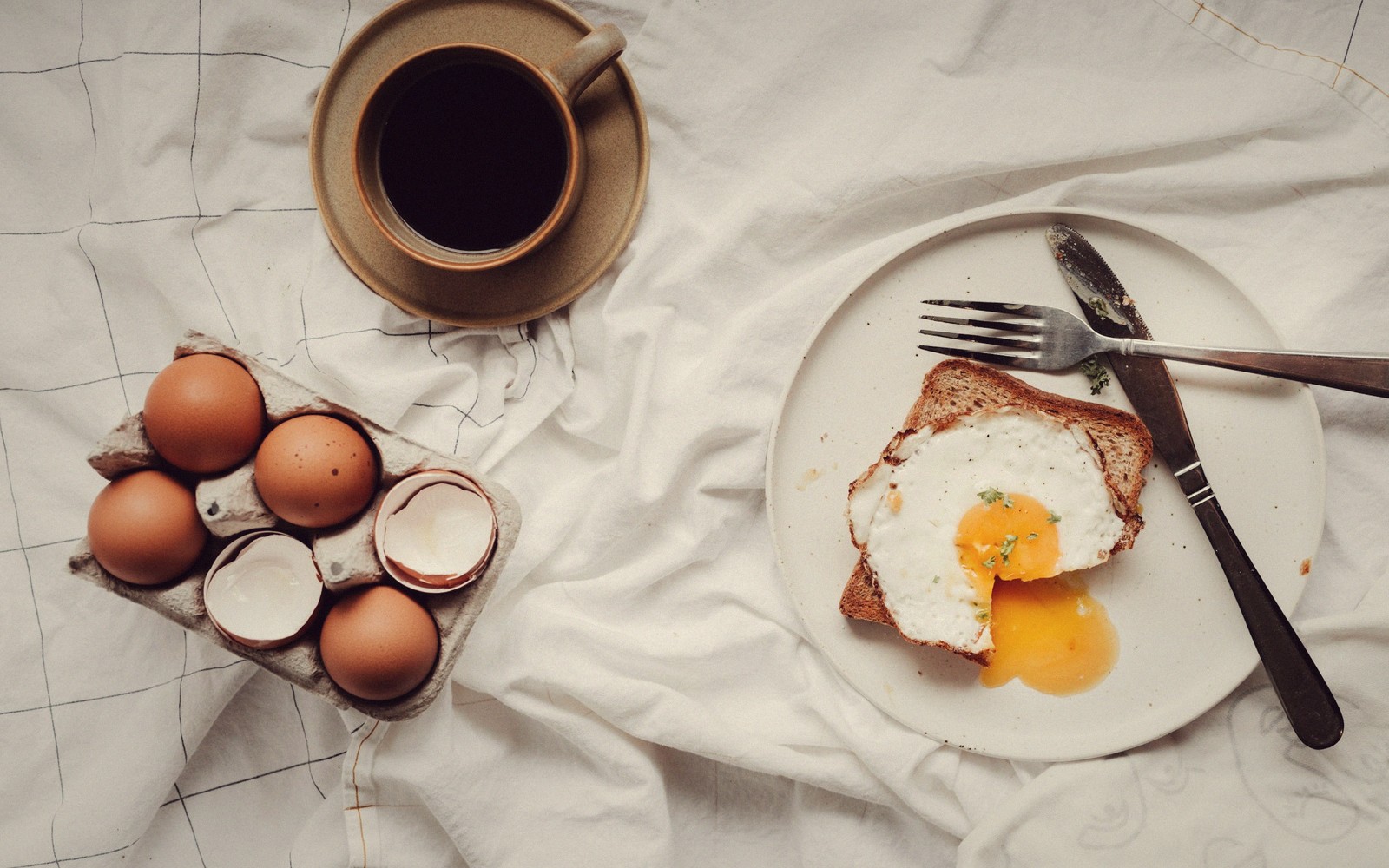 Fried egg on toast beside a cracked egg carton and cup of coffee