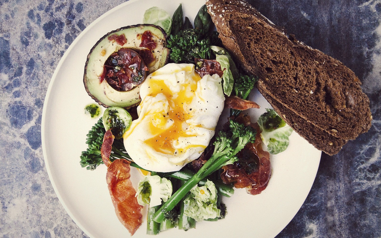 Poached egg with bacon, broccolini, stuffed avocado and dark rye bread on a plate