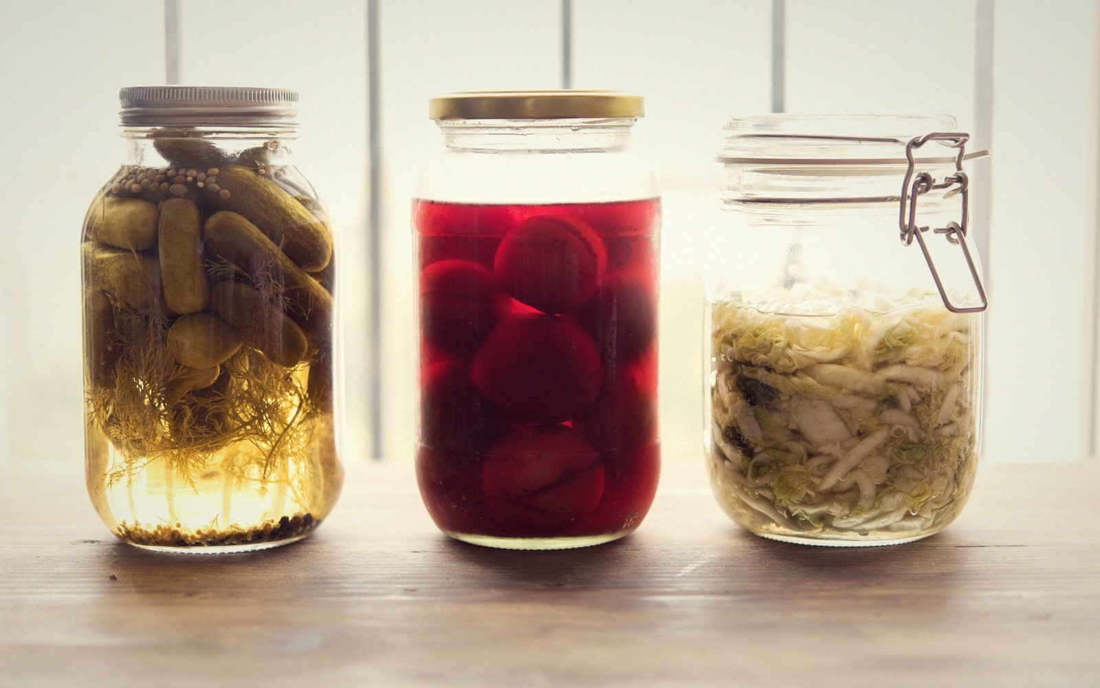 Three glass jars on a wooden table holding pickled cucumbers, beets, and sauerkraut