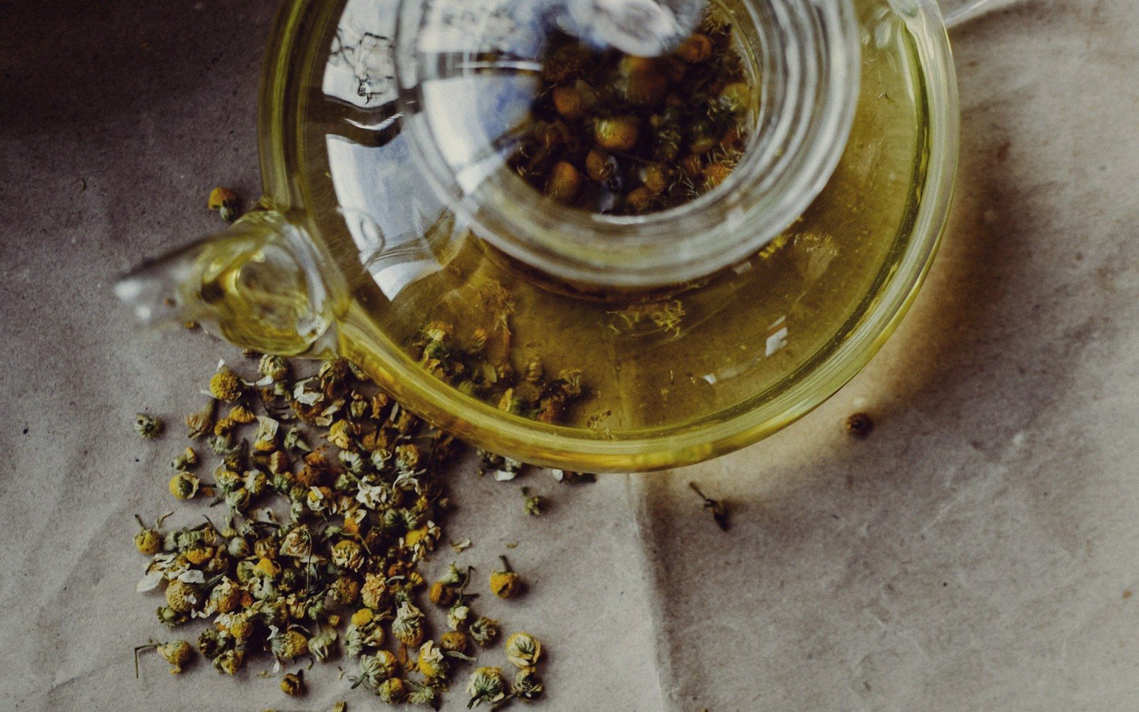 Glass teapot brewing chamomile flowers, surrounded by scattered dried chamomile buds