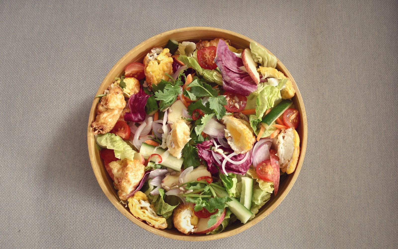 A wooden bowl of salad with tomatoes, cucumber, red onion, herbs and fried chicken pieces