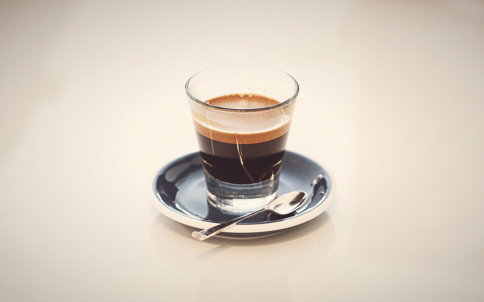 Espresso in a small glass cup with crema, resting on a dark saucer with a spoon